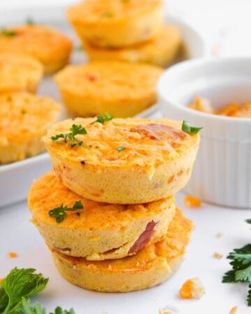 Three Ham and Cheese Egg Bites stacked on each other, garnished with parsley. More egg muffins are visible in the background on a plate, along with scattered cheese and parsley on a white surface.