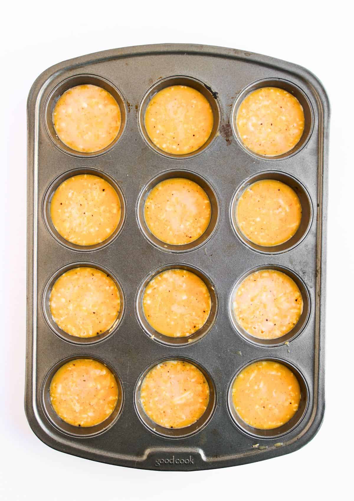 A muffin tin with twelve wells, each filled with an orange batter containing visible flecks, ready to be baked.
