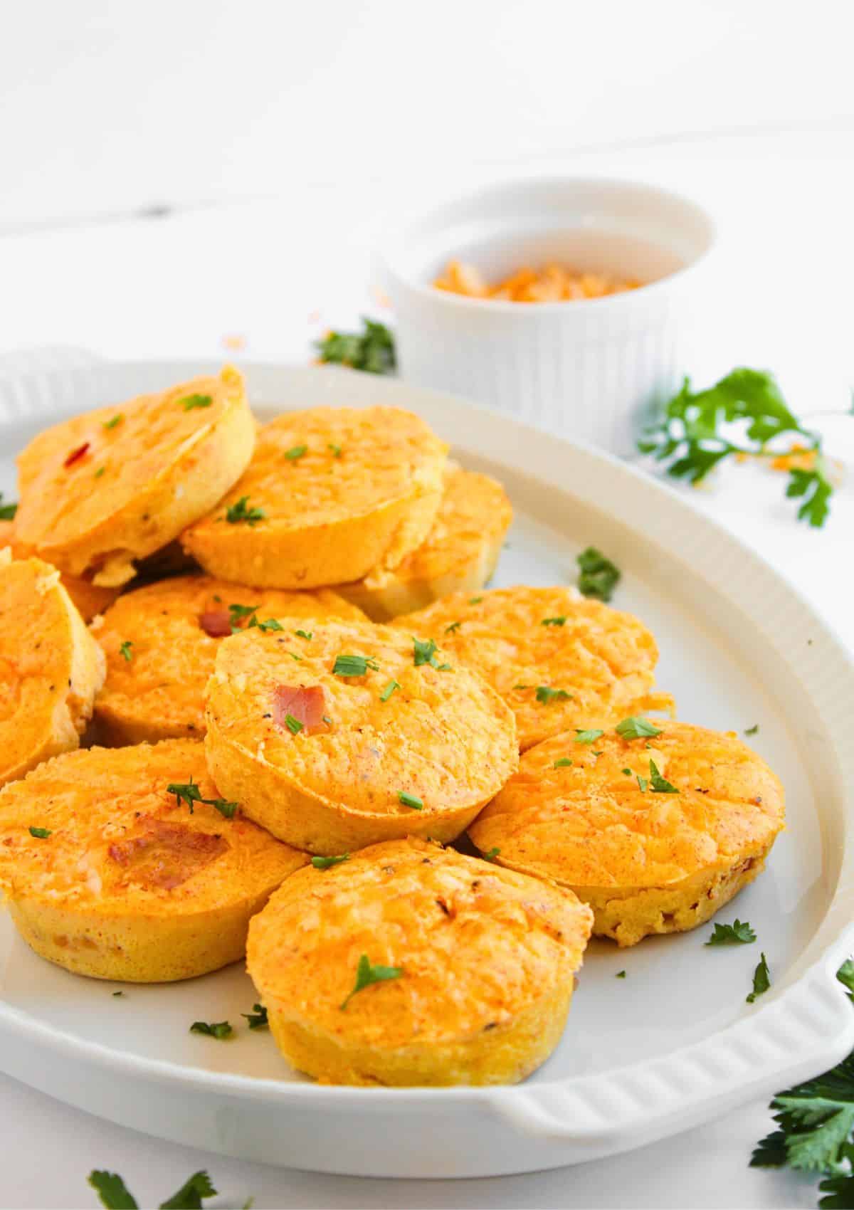 A white plate with several mini egg muffins, reminiscent of Ham and Cheese Egg Bites, garnished with chopped herbs. More herbs and a small white ramekin with grated cheese are in the background on a white surface.