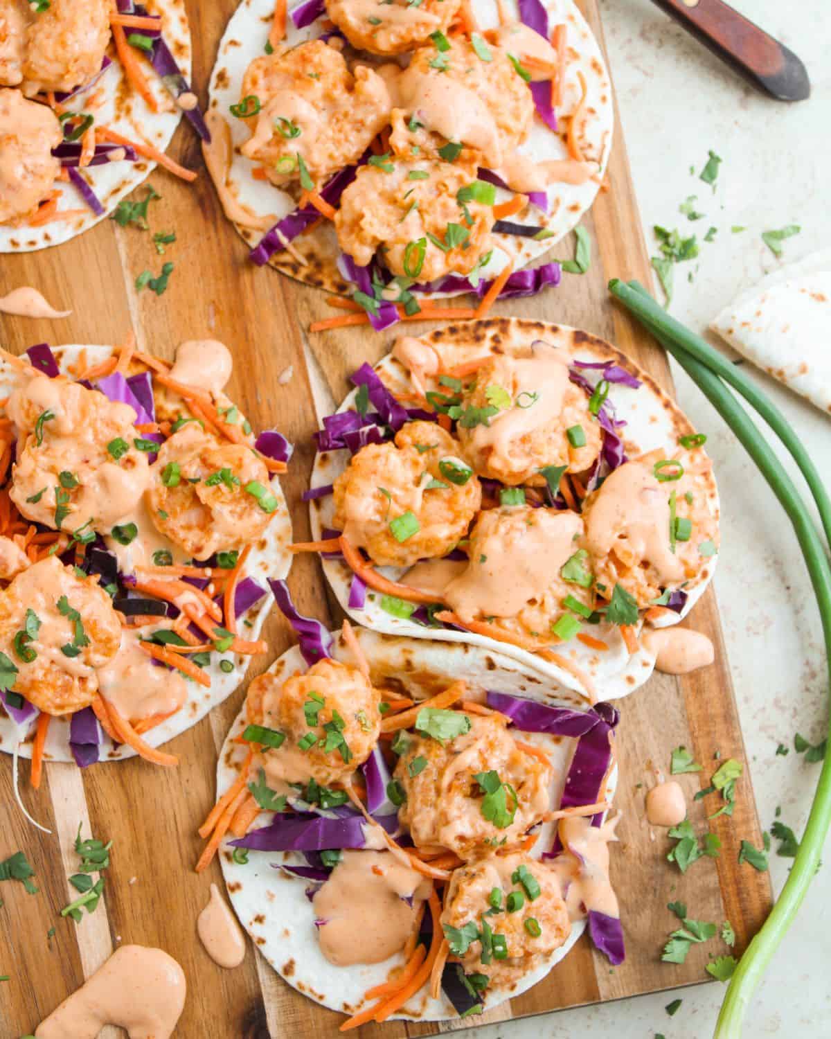 Four Bang Bang Shrimp Tacos on tortillas topped with purple cabbage, shredded carrots, chopped herbs, and drizzled with creamy orange sauce, arranged on a wooden board with green onions nearby.