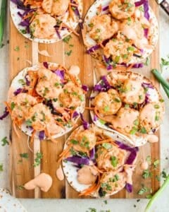 Six small Bang Bang Shrimp Tacos on a wooden board, filled with shrimp, shredded purple cabbage, carrots, and drizzled with a creamy orange sauce, garnished with chopped herbs. Scattered ingredients and sauce drops surround the tacos.