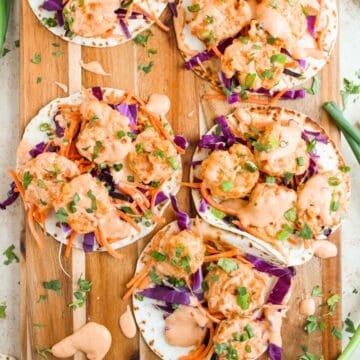 Six small Bang Bang Shrimp Tacos on a wooden board, filled with shrimp, shredded purple cabbage, carrots, and drizzled with a creamy orange sauce, garnished with chopped herbs. Scattered ingredients and sauce drops surround the tacos.