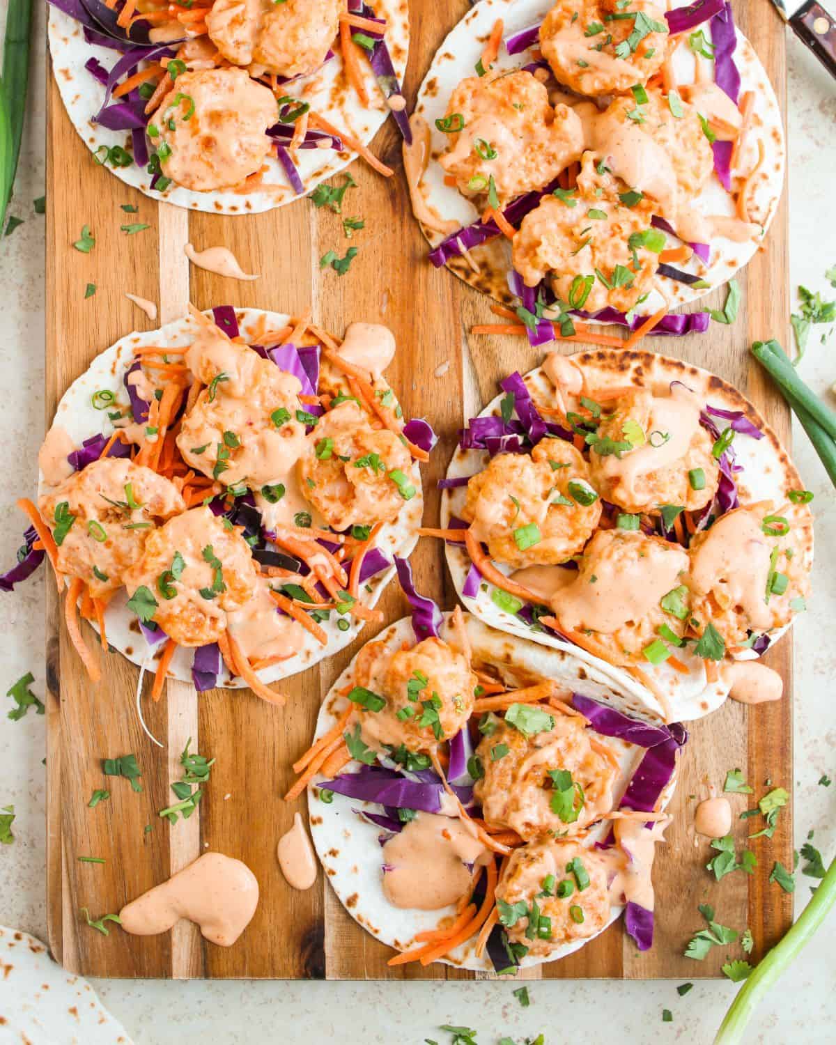 Six small Bang Bang Shrimp Tacos on a wooden board, filled with shrimp, shredded purple cabbage, carrots, and drizzled with a creamy orange sauce, garnished with chopped herbs. Scattered ingredients and sauce drops surround the tacos.