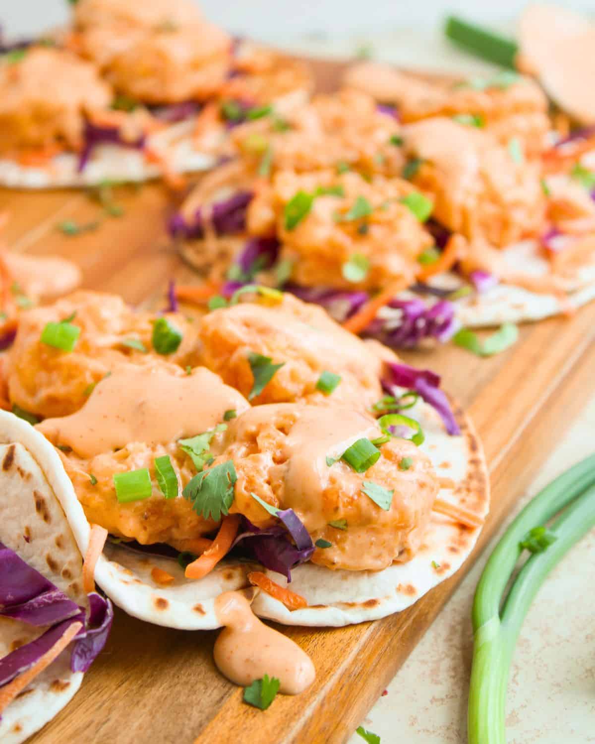 Enjoy Bang Bang Shrimp Tacos—soft tortillas loaded with crispy breaded shrimp, shredded purple cabbage, carrots, green onions, cilantro, and a creamy orange sauce, all artfully served on a wooden board.