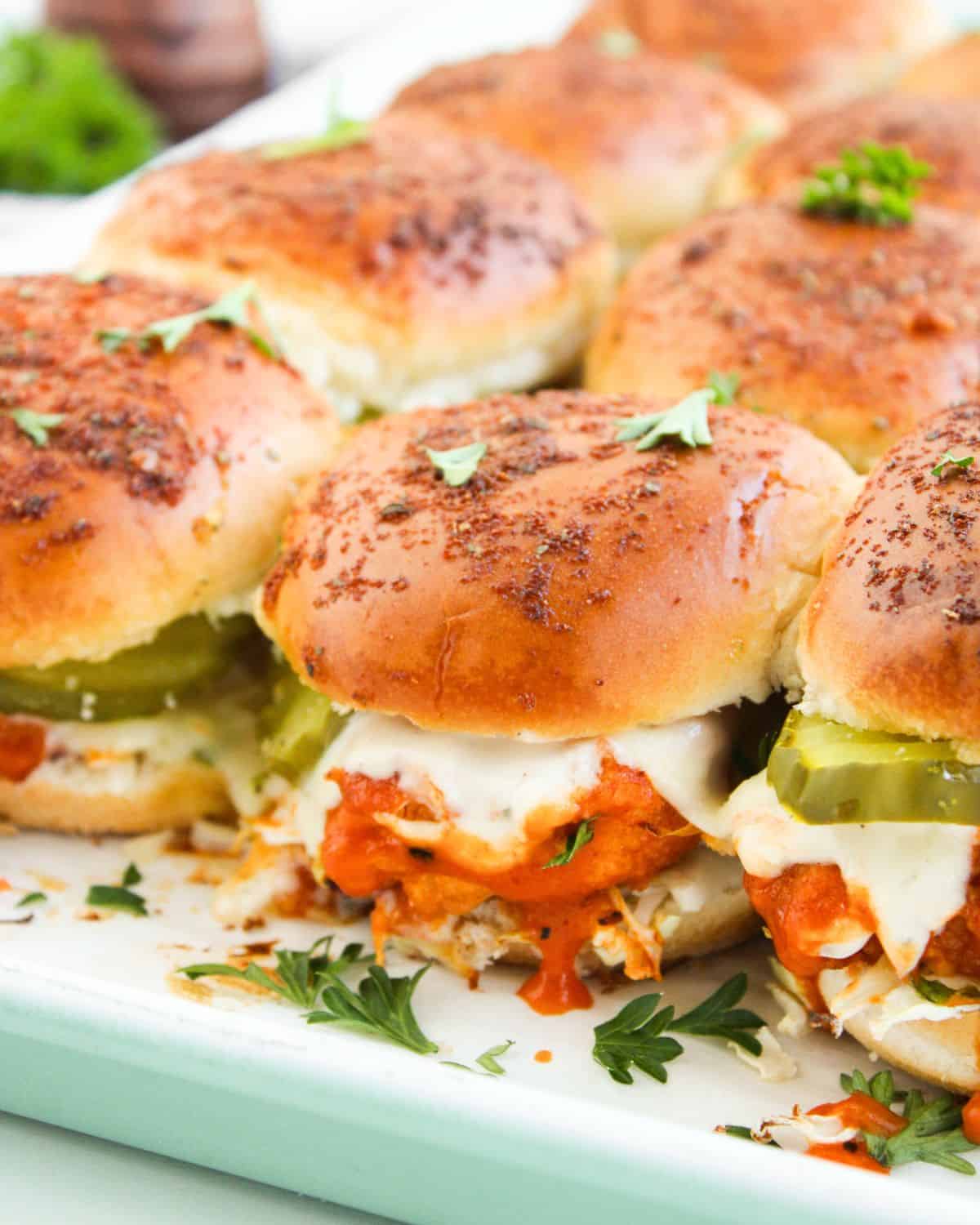 A close-up of several Buffalo Chicken Sliders with golden buns, melted cheese, shredded meat in a red sauce, sliced pickles, and fresh herbs on a light-colored tray.