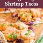 Three Bang Bang Shrimp Tacos topped with creamy sauce, chopped green onions, and purple cabbage, served on a wooden board. Text above reads “Mama Maggie’s Kitchen Bang Bang Shrimp Tacos.”.