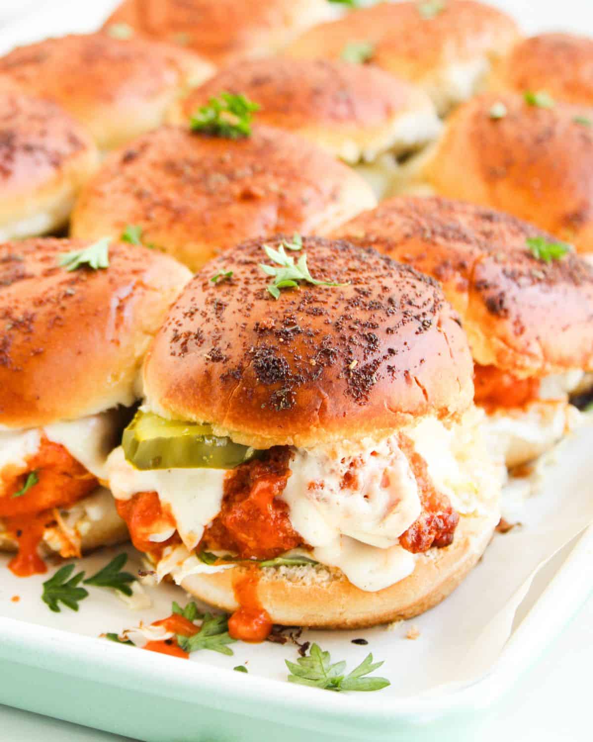 A tray of Buffalo Chicken Sliders with browned buns, melted cheese, chicken in tomato sauce, and pickle slices, garnished with fresh parsley.
