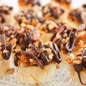Easy Frozen Banana Snickers Bites: Sliced bananas topped with caramel, chopped nuts, and drizzled with chocolate, all arranged beautifully on a glass plate.