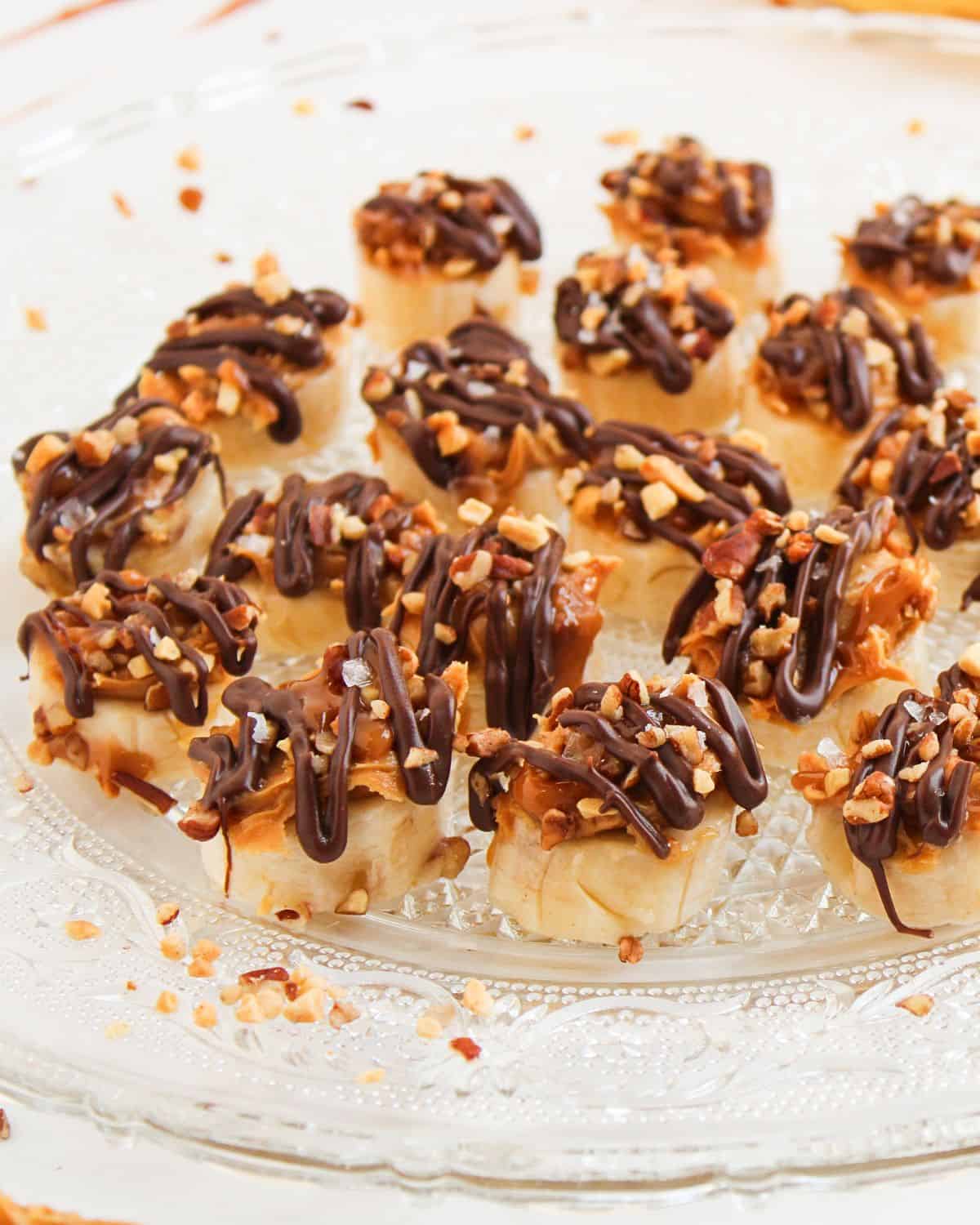 Easy Frozen Banana Snickers Bites are small, bite-sized desserts topped with chopped nuts and drizzled with chocolate, arranged on a clear glass plate. Each treat sits on a pastry or cracker base for a delightful crunch.