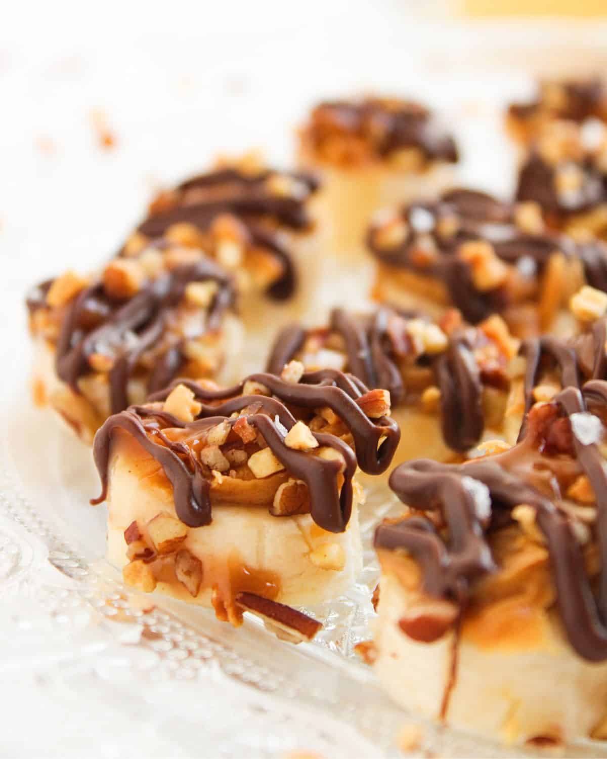 Close-up of Easy Frozen Banana Snickers Bites—banana slices topped with chopped nuts and drizzled with melted chocolate, arranged on a clear glass plate.
