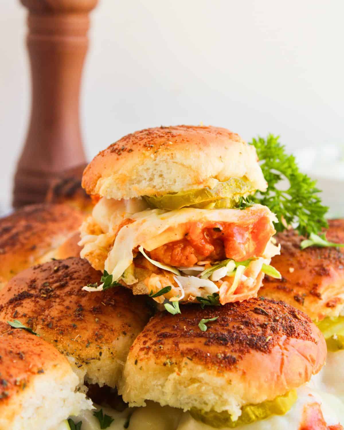 A stack of golden-brown Buffalo Chicken Sliders filled with fried chicken, pickles, and coleslaw, garnished with fresh parsley, with a pepper mill blurred in the background.