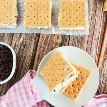 Two Mascarpone Ice Cream Sandwiches with graham crackers sit on a white plate next to a pink checkered cloth. More sandwiches rest on parchment paper above, with a bowl of chocolate chips on the wooden table nearby.