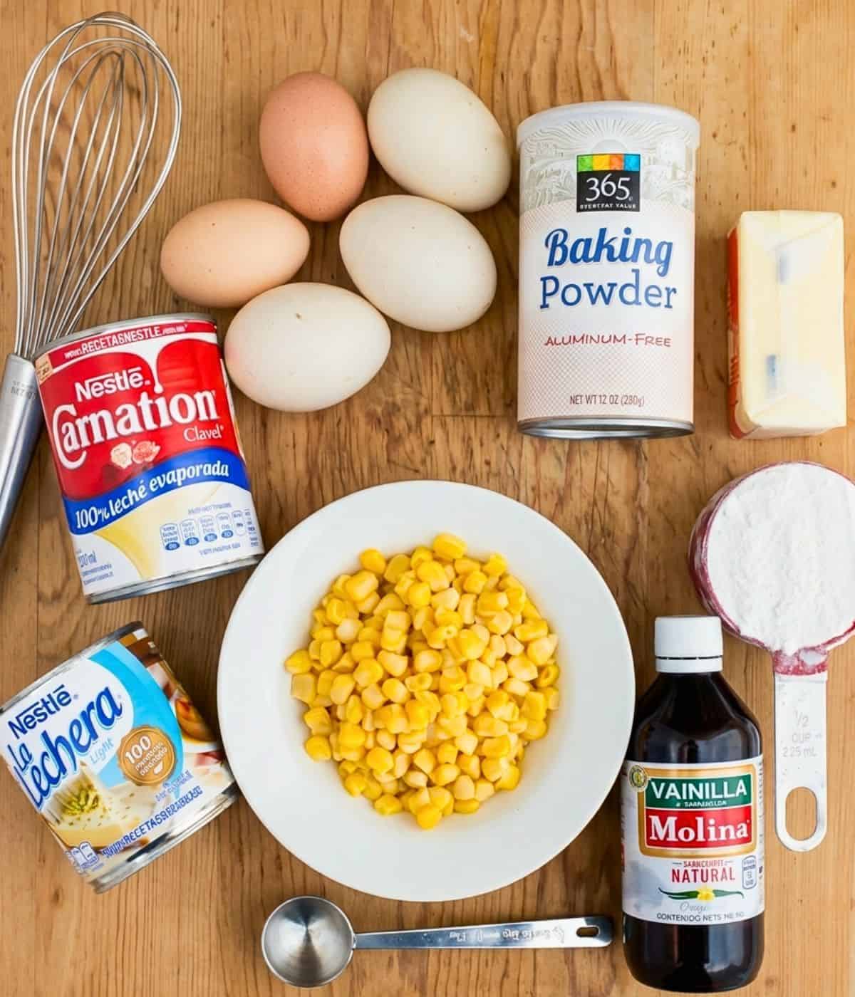 Various baking ingredients displayed on a wooden surface, including eggs, butter, baking powder, evaporated milk, sweetened condensed milk, flour, corn kernels, vanilla extract, a whisk, and a measuring spoon.