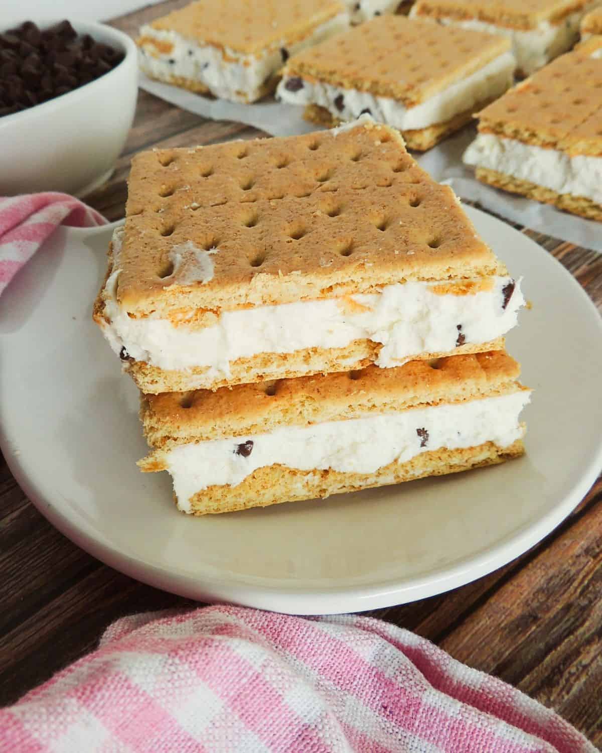 Two Mascarpone Ice Cream Sandwiches with graham crackers sit on a white plate next to a pink checkered cloth. More sandwiches rest on parchment paper above, with a bowl of chocolate chips on the wooden table nearby.