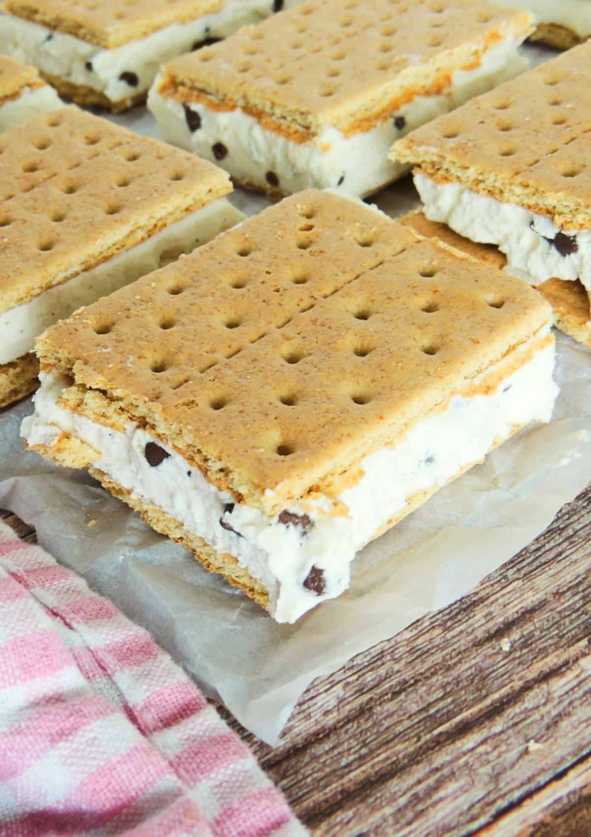 Two Mascarpone Ice Cream Sandwiches with graham crackers sit on a white plate beside a pink checkered cloth, while more sandwiches rest on parchment paper above, and a bowl of chocolate chips sits on the wooden table nearby.