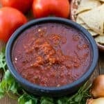 A black bowl of chunky red salsa, made from an authentic Chipotle salsa recipe, sits on a table with fresh tomatoes, cilantro, an onion, and tortilla chips nearby.