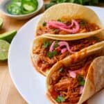 Three corn tortillas filled with Cochinita Pibil-style shredded, saucy chicken, topped with pickled red onions and cilantro, served on a white plate. Slices of lime, jalapeños, pickled onions, and fresh cilantro are in the background.