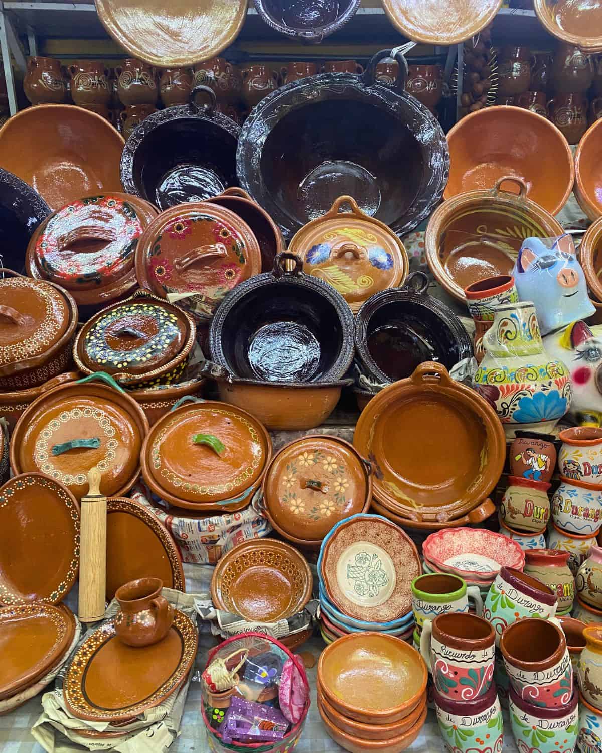 A market stall displays various traditional Mexican clay dishes, pots, bowls, and mugs, some plain brown and others decorated with colorful floral and geometric designs.