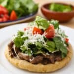 A Mexican sope topped with grilled beef, chopped lettuce, diced tomatoes, and shredded cheese is served on a white plate. In the background are fresh lettuce, tomatoes, and a bowl of green sauce—classic flavors of Mexican sopes.