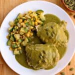 A white plate with two pieces of chicken covered in vibrant Pipian Verde sauce and pumpkin seeds, served with sautéed zucchini and corn. A bowl of pumpkin seeds is nearby on a wooden surface.