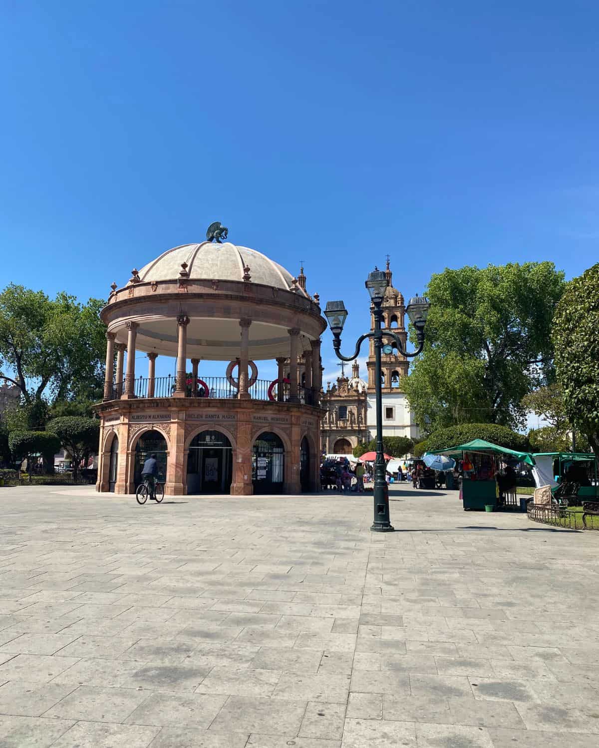 A round, ornate gazebo stands in a sunny plaza with trees, a cathedral tower in Durango, Mexico in the background, and people walking or sitting nearby under a clear blue sky.