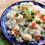 A blue plate of cooked Arroz Blanco mixed with green peas and diced carrots, garnished with fresh cilantro. A fork and cilantro leaves are visible in the background.