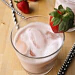 A glass of pink iced strawberry agua fresca garnished with a fresh strawberry, with more strawberries and two black-and-white striped straws on a wooden surface.