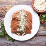 A white plate with enfrijoladas—enchiladas smothered in rich refried bean sauce—topped with crumbled cheese and fresh cilantro. Next to the plate are bowls of cheese, sliced jalapeños, fresh cilantro, and pinto beans on a rustic wooden table.