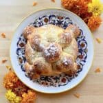 Round loaf of Pan de Muerto dusted with sugar on a decorative plate, surrounded by vibrant orange and yellow marigold flowers on a light wooden surface.