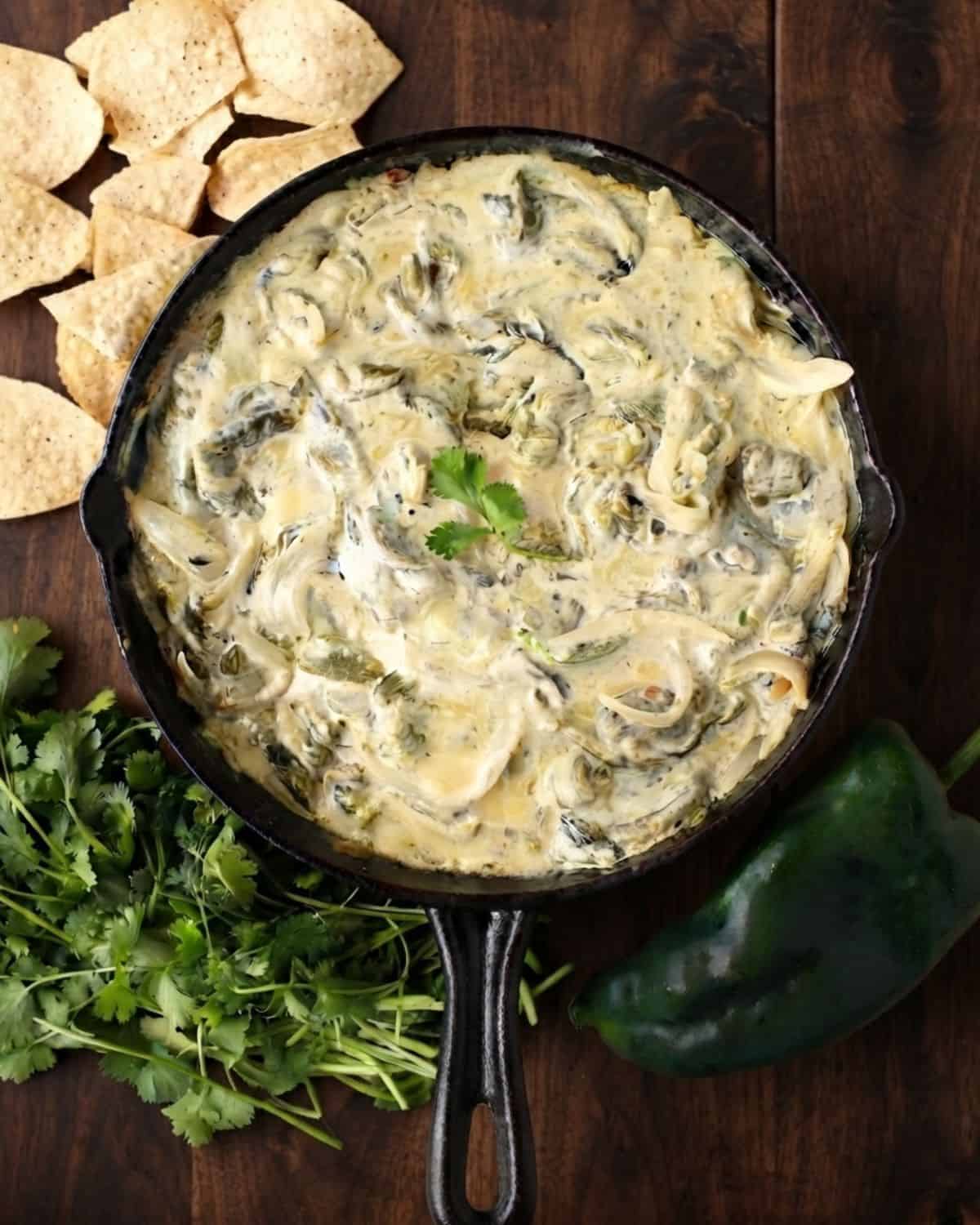 A skillet filled with creamy poblano dip, inspired by classic Rajas con Crema, garnished with cilantro and surrounded by tortilla chips, fresh cilantro, and a whole poblano pepper on a dark wooden surface.