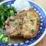 A plate with a cooked pork chop topped with sauce and herbs, served with white rice, broccoli, and sliced jalapeño on a blue patterned dish—this vibrant meal is reminiscent of chuletas en salsa verde.