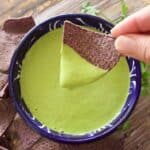 A hand dips a blue corn tortilla chip into a bowl of creamy green Creamy Jalapeno Salsa, surrounded by more chips on a wooden surface.