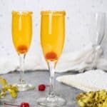 Two champagne flutes with Golden Champagne Cocktail, sugared rims, and cherries at the bottom. A cherry, gold ribbon, and napkin are nearby, with a white cloth and empty glass in the background.