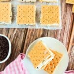 Eight Mascarpone Ice Cream Sandwiches with chocolate chips on graham crackers are arranged on parchment. Two are on a white plate with a pink cloth, and a bowl of chocolate chips sits nearby on a wooden table.