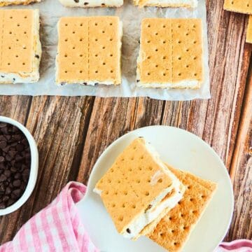 Eight Mascarpone Ice Cream Sandwiches with chocolate chips on graham crackers are arranged on parchment. Two are on a white plate with a pink cloth, and a bowl of chocolate chips sits nearby on a wooden table.