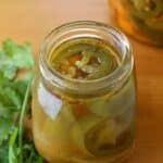 A glass jar filled with chiles en vinagre—pickled jalapeño slices in brine—sits on a wooden surface next to fresh cilantro leaves.