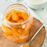 A glass jar filled with sliced Mexican Pickled Carrots sits on a wooden board, with a spoon holding carrot slices beside it. A patterned bowl is in the background.