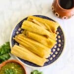 A plate of Tamales de Rajas sits on a blue patterned dish, surrounded by green salsa, fresh cilantro, and a clay cup filled with a dark drink on a white marble surface.