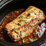 A Slow Cooker Honey Lime Chipotle Pork Loin topped with herbs and sesame seeds sits in sauce inside a black slow cooker.