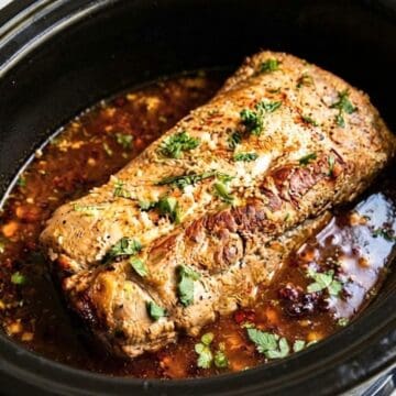 A Slow Cooker Honey Lime Chipotle Pork Loin topped with herbs and sesame seeds sits in sauce inside a black slow cooker.
