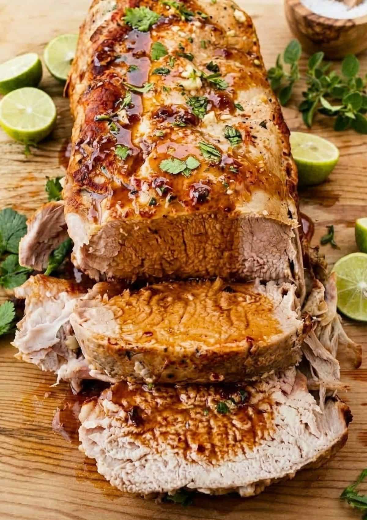 Sliced Slow Cooker Honey Lime Pork Loin with glaze, garnished with chopped herbs, surrounded by lime halves and fresh herbs on a wooden cutting board.