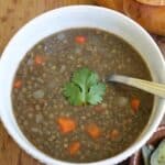 A bowl of Sopa de Lentejas with diced carrots and onions, topped with a cilantro leaf, and a spoon resting inside. Bread is visible in the background.