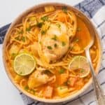 A bowl of Sopa de Pollo con Fideos with noodles, vegetables, lime slices, and fresh herbs. A spoon rests in the bowl, which is set on a blue and white checkered cloth.