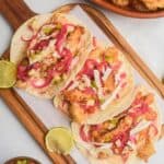 Three Tacos de Pescado on a wooden board, topped with pickled onions, shredded cabbage, and salsa. Two lime wedges are on the side. A bowl of salsa and a plate of fried fish are nearby.