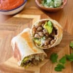 A Carnitas Burrito cut in half on a wooden board, filled with meat, avocado, lettuce, and onions. Salsa and a bowl of chopped onions are in the background. Fresh cilantro leaves are scattered nearby.